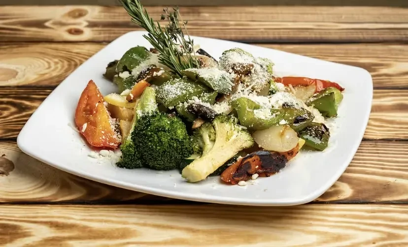 Plate of roasted vegetables with cheese, rosemary, and herbs on wood.