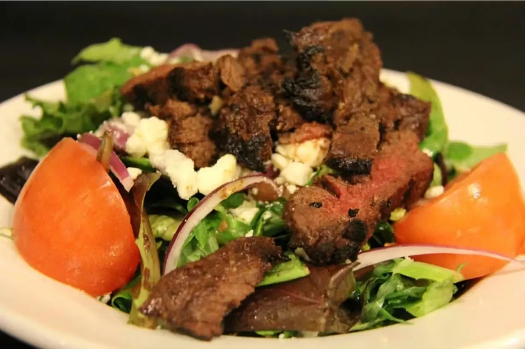 Close-up steak salad with mixed greens, grilled steak, crumbled cheese, and tomatoes.