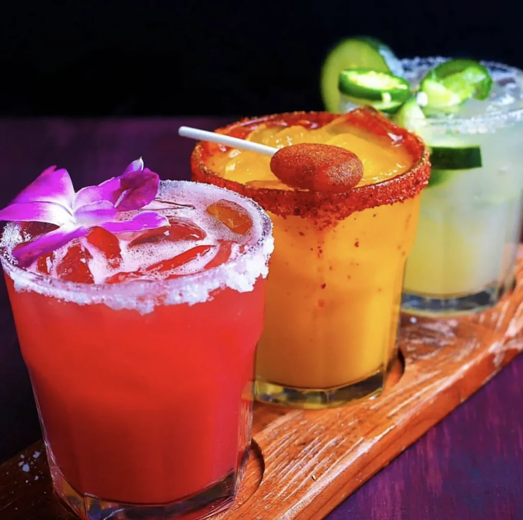 Three colorful cocktails with unique garnishes on a wooden tray.