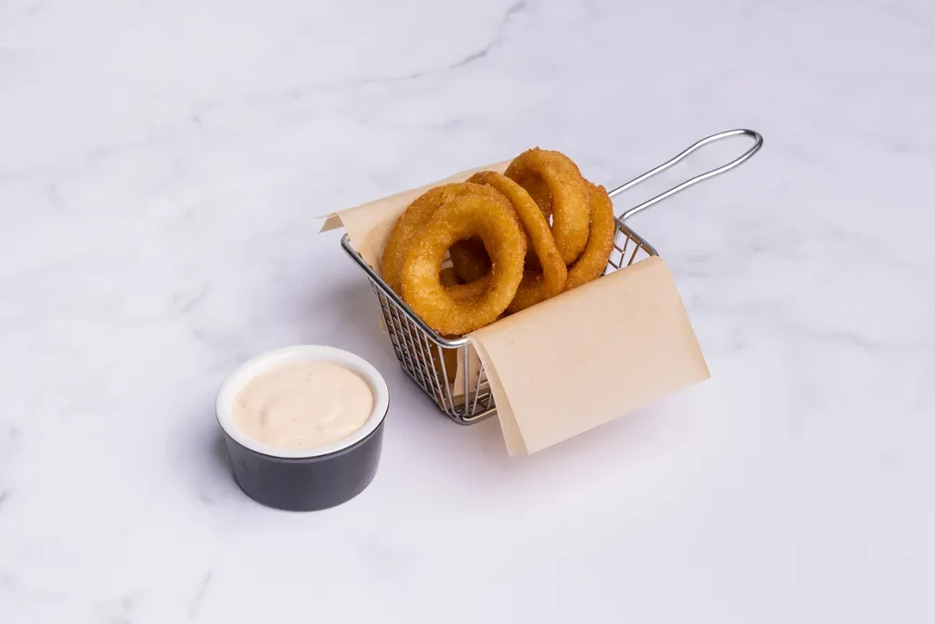 Golden-brown onion rings in frying basket with dipping sauce on marble.