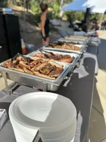 Outdoor buffet with chafing dishes of barbecue meats and disposable plates.