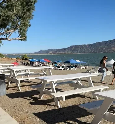 Sunny outdoor gathering by a lake with picnic tables and colorful tents.