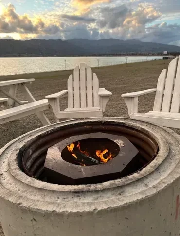 Burning fire pit, Adirondack chairs, lake, mountains under twilight sky.