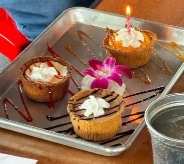 Four mini desserts, one with a lit candle, on a decorated silver tray.