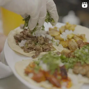 White-gloved hand sprinkles fresh cilantro onto meat tacos.