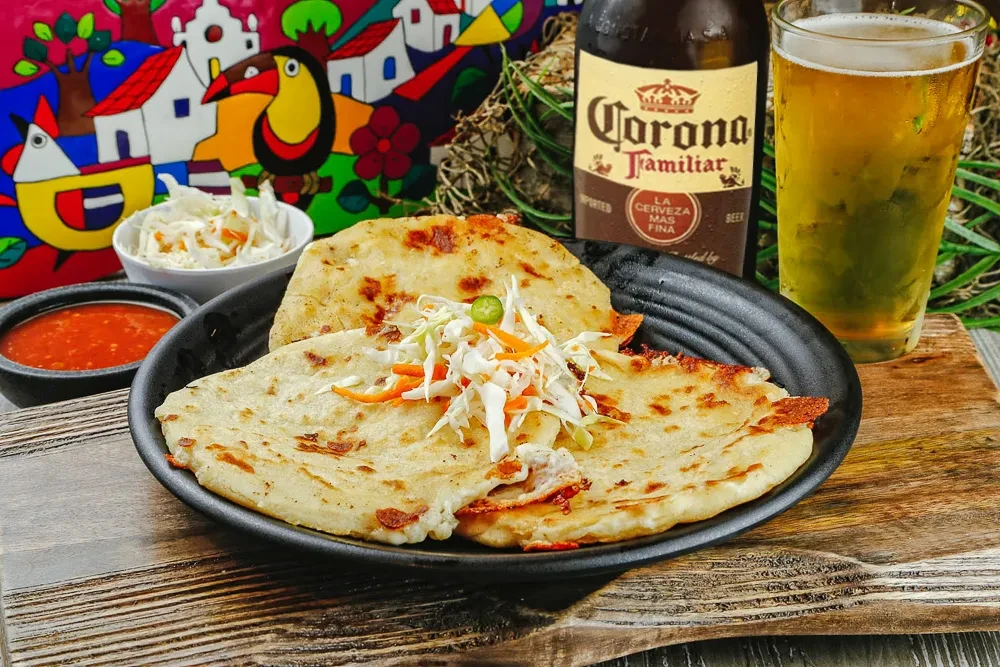 Three pupusas topped with curtido, red salsa, beer, and folk art.