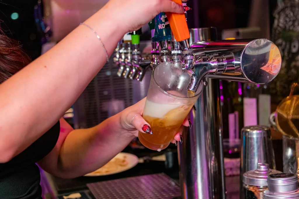 Hands pouring foamy beer from a tap in a neon-lit bar.