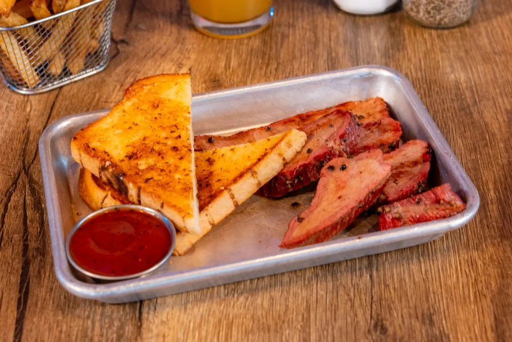 Sliced smoked meat, toast, red sauce, and fries on a metal tray.