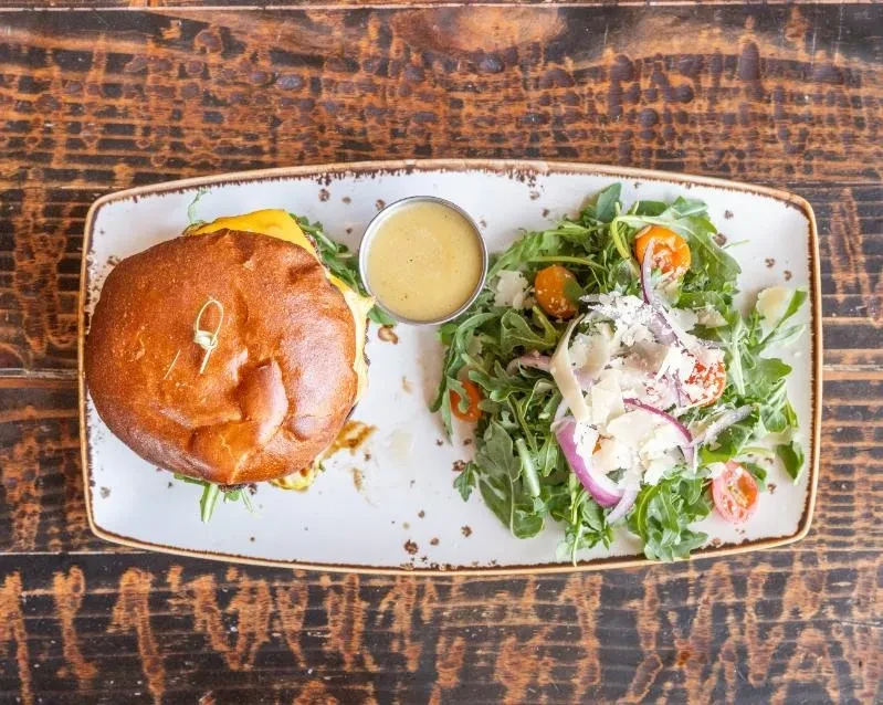 Golden-brown burger, arugula salad, and dressing cup on white plate.