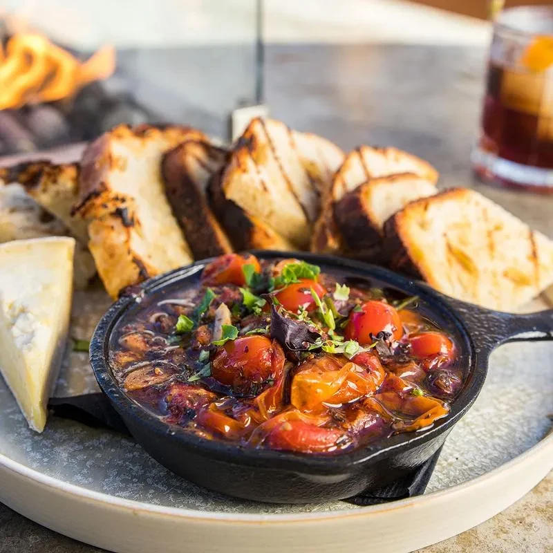 Cast-iron skillet with rustic stew, bread, cheese, and outdoor fire.