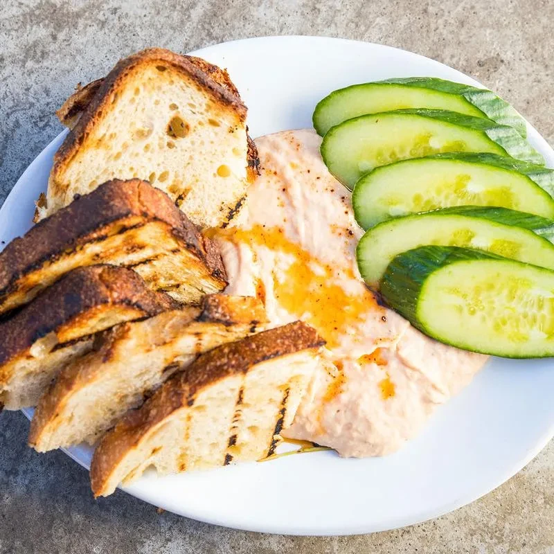 Grilled bread, pink dip with oil, and fanned cucumber on a plate.