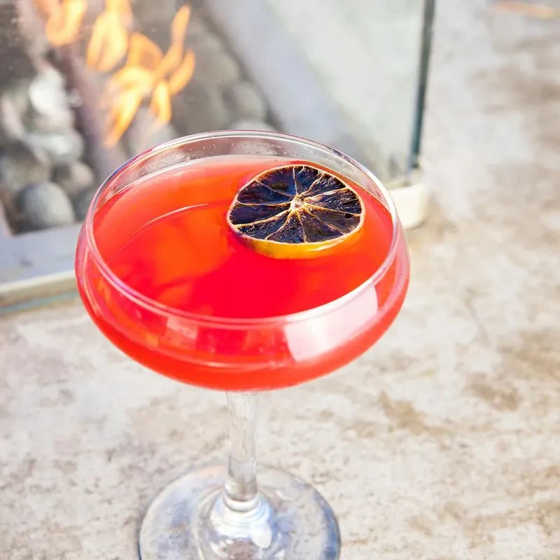 Red cocktail in coupe glass with dried citrus, fire pit background.