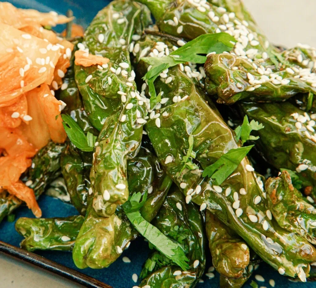 Cooked green peppers, herbs, sesame, and kimchi on a dark plate.