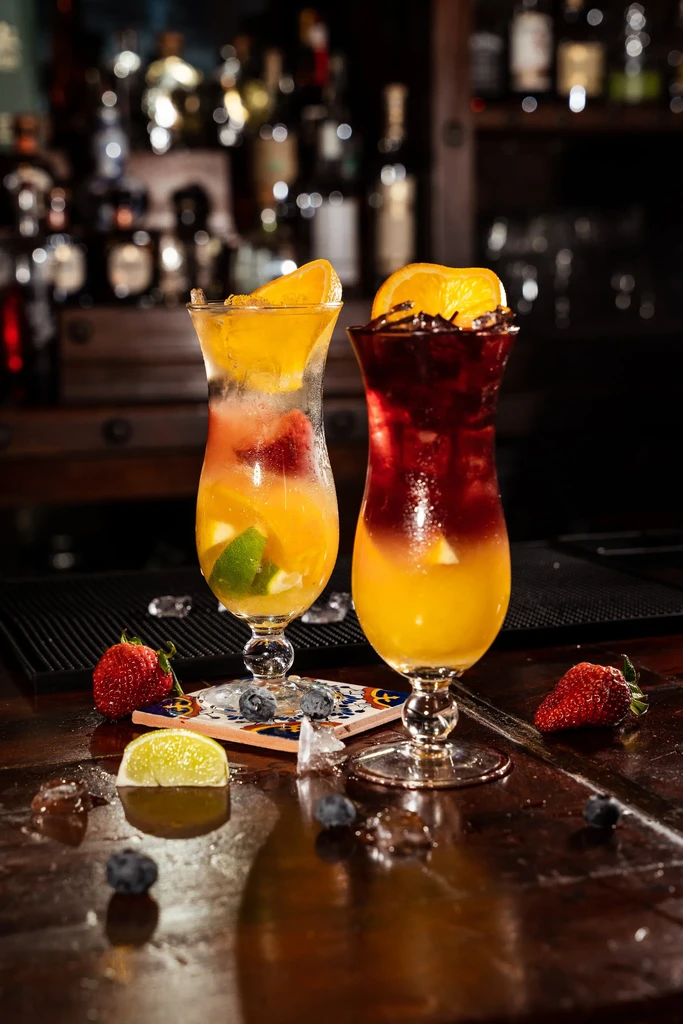 Two colorful cocktails with fruit and ice on a wooden bar.