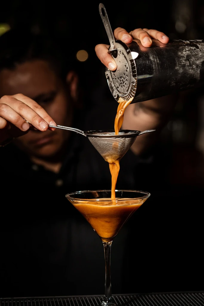 Bartender double-straining an amber cocktail into a martini glass.