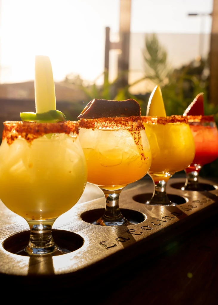 Four vibrant, spice-rimmed beverages with unique garnishes on a wooden tray.