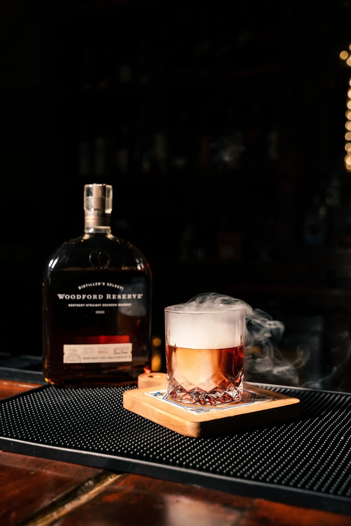 Faceted glass smoked cocktail beside Woodford Reserve bourbon bottle.