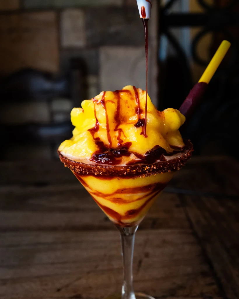 Yellow slushie in martini glass, drizzled with dark sauce, chili-lime rim.
