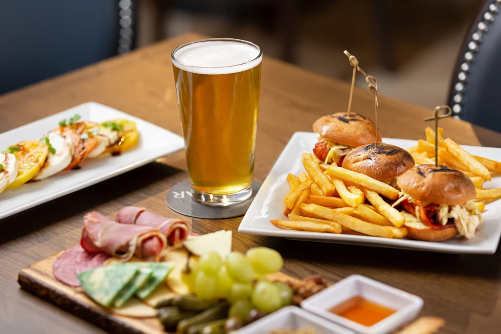 A wooden table with beer, sliders, Caprese salad, and charcuterie board.