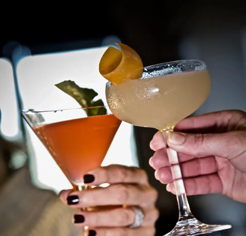 Two cocktails, orange and beige, clinking in a toast.