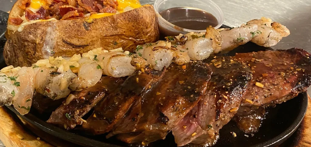 Sliced steak, shrimp, loaded baked potato on a sizzling plate.