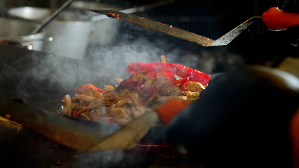 Sizzling chicken and vegetables stirred by gloved hands.