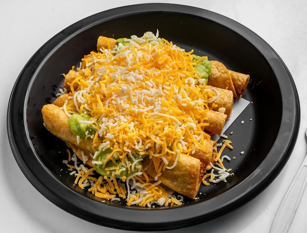 Golden-brown taquitos with shredded cheese and green sauce on a black plate.
