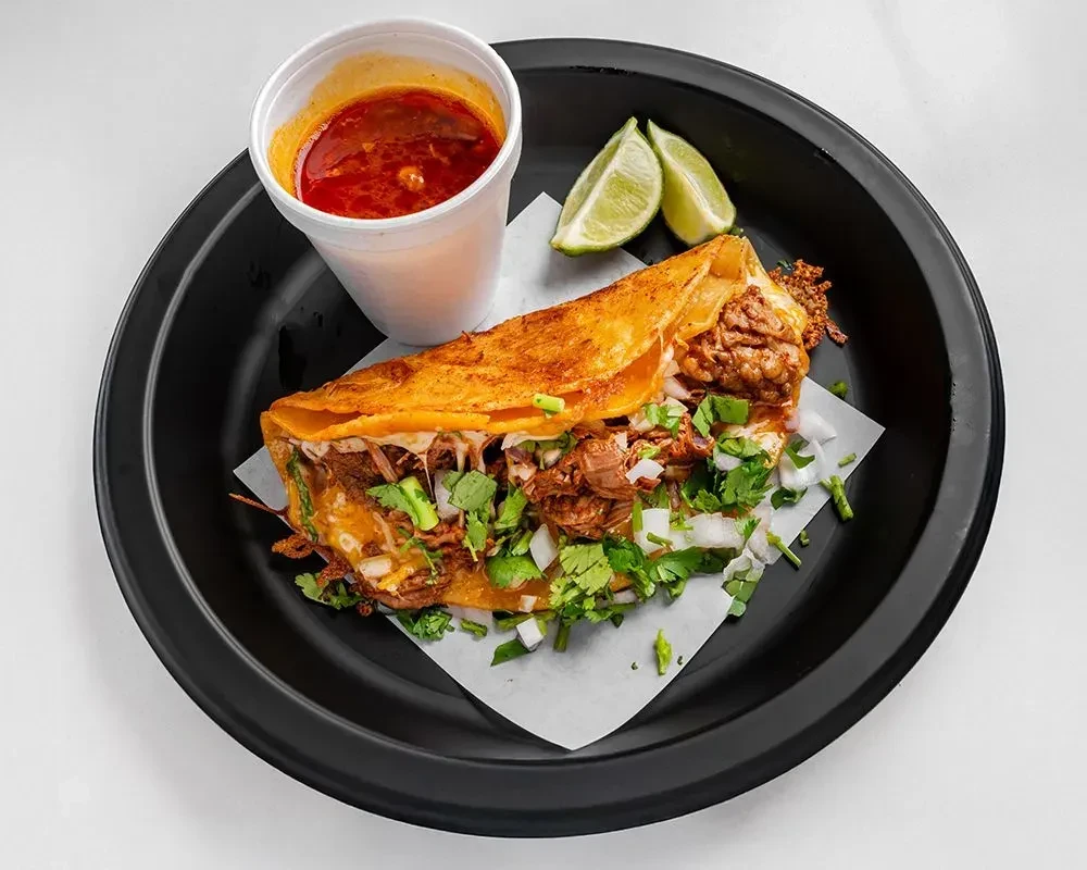 Birria taco filled with meat, cheese, consommé, and lime wedges.