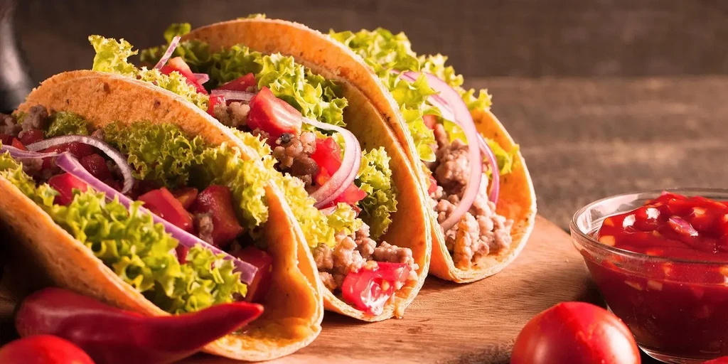 Three tacos with meat and vegetables on a cutting board with salsa.