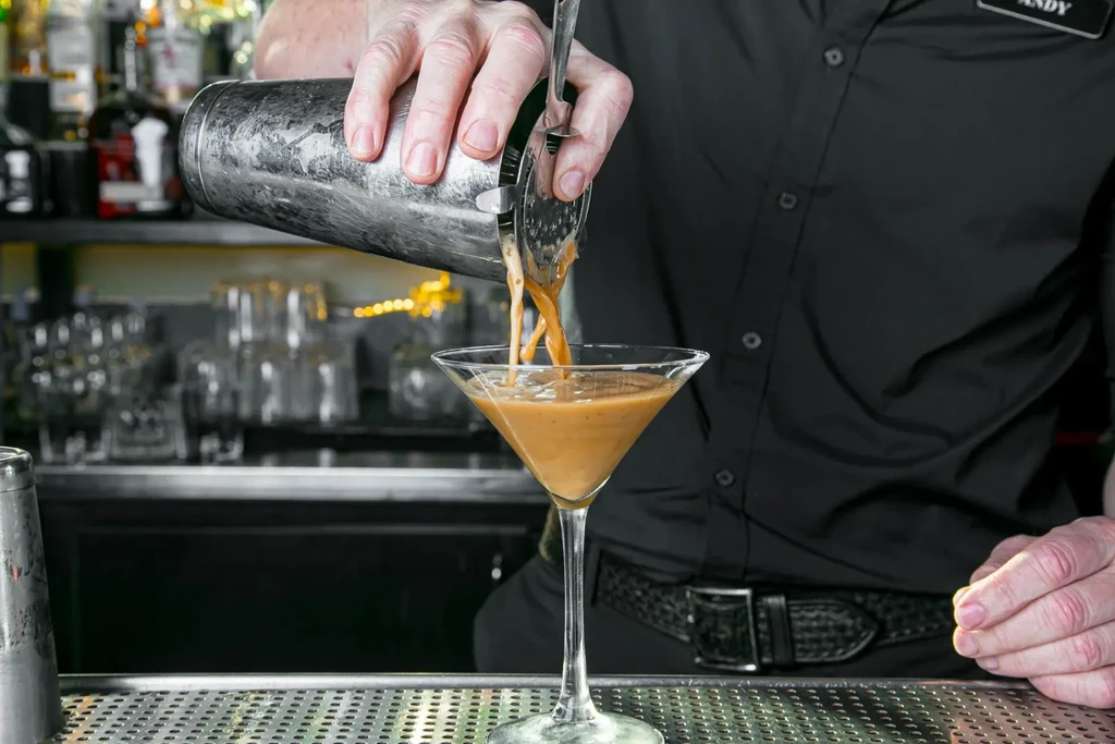 A bartender's hands pour a creamy cocktail into a martini glass.