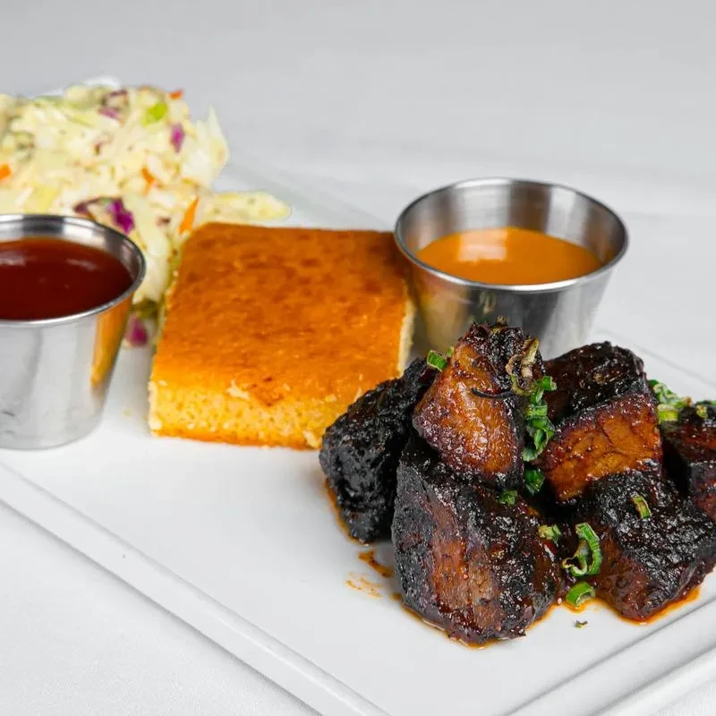 Barbecued meat, cornbread, coleslaw, and two sauces on a white plate.