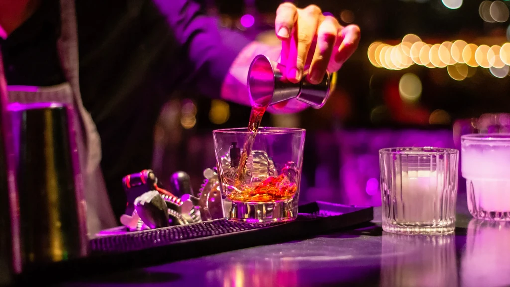 Bartender's hands pour golden drink into glass in purple-lit bar.