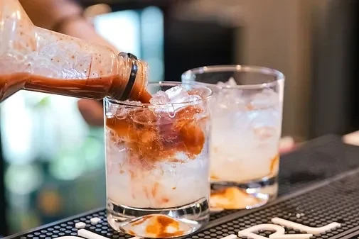 Hand pouring reddish-brown liquid into iced glass, creating swirl.