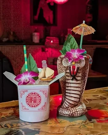 Two ornate cocktails in a Chinese take-out box and cobra tiki mug.