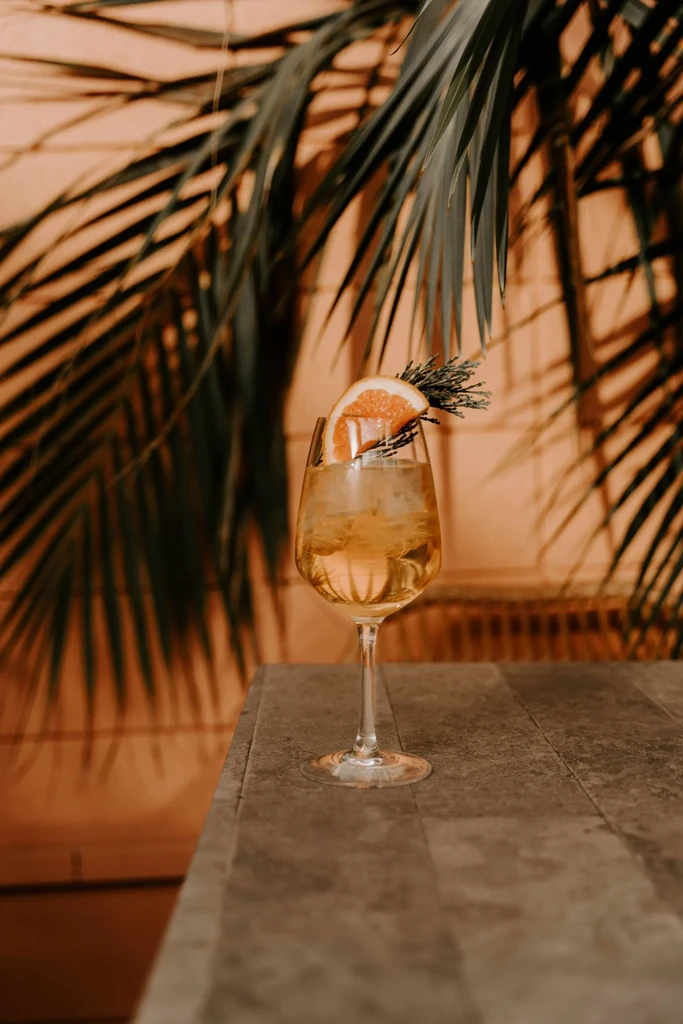 Golden cocktail with ice, citrus, and rosemary against palm frond background.