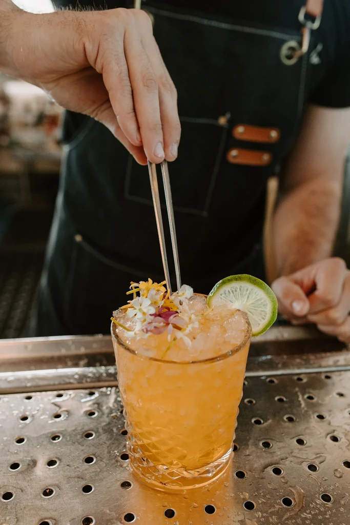 Bartender meticulously garnishes an orange cocktail with lime and colorful flowers.