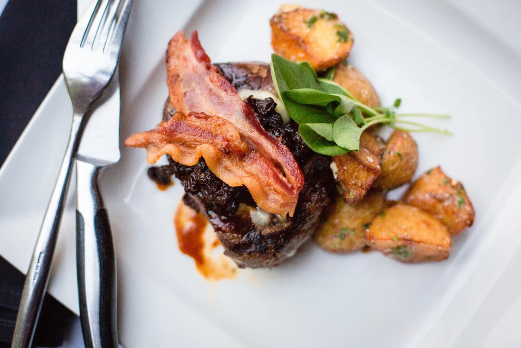 Plated grilled patty, bacon, melted topping, roasted potatoes, sauce, and herbs.