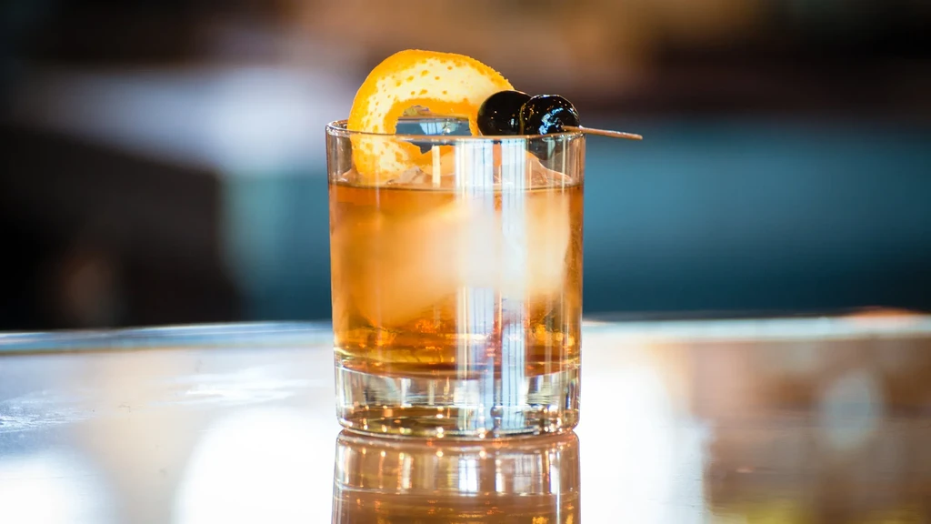 Amber-brown cocktail with ice, orange peel, and cherries on a reflective surface.