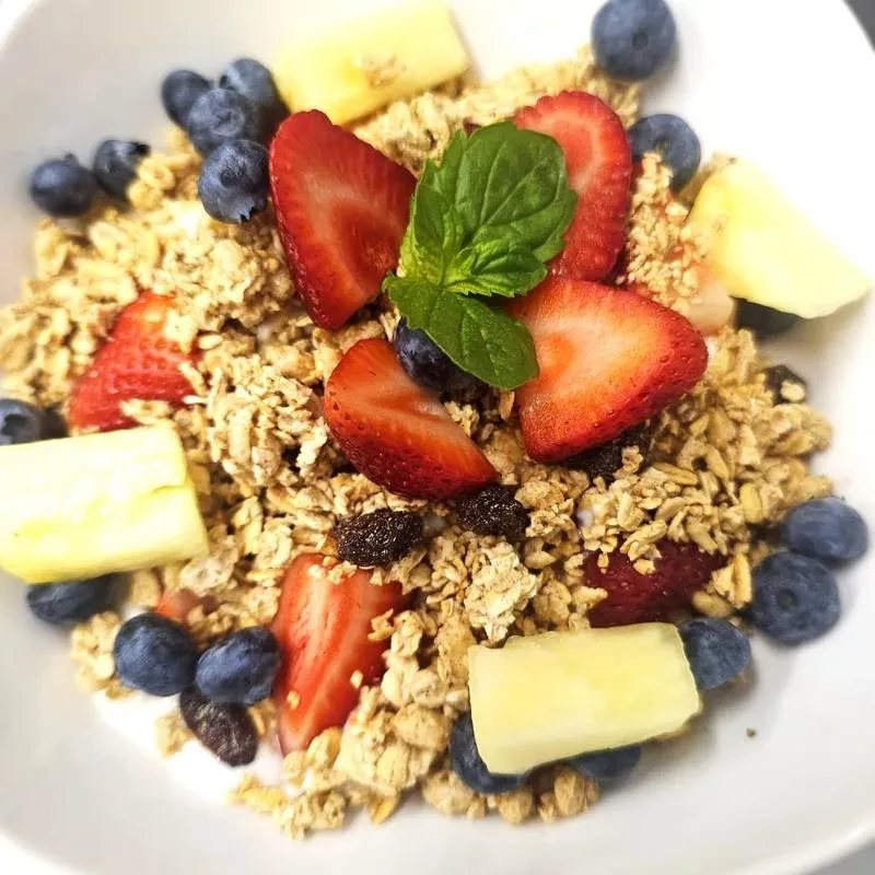 Close-up of granola with berries, pineapple, raisins, and mint in a white bowl.
