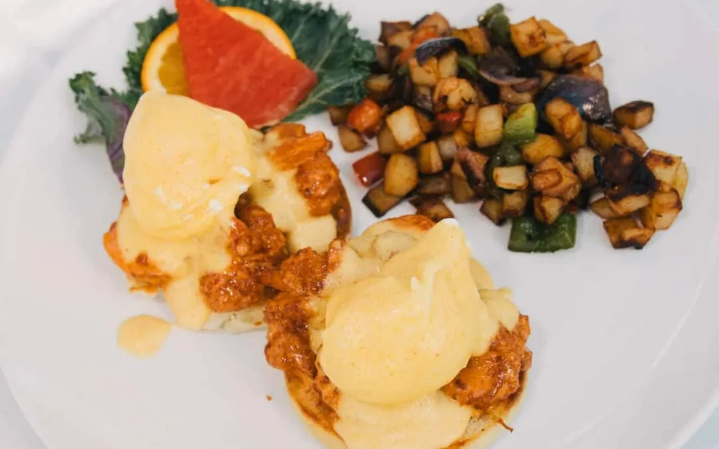 Plated eggs benedict with potatoes, kale, orange, and red fruit.