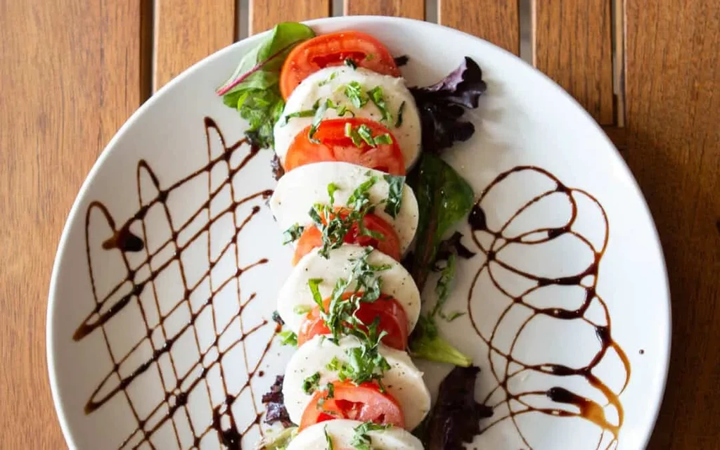 Overhead Caprese salad on white plate: mozzarella, tomatoes, basil, balsamic.