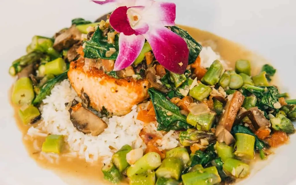 Seared salmon on rice with vegetables, sauce, and an orchid.