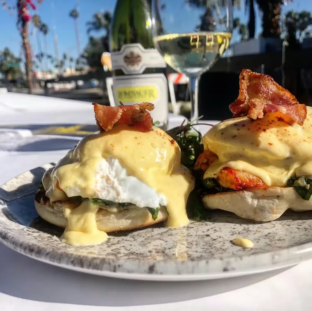 Two Eggs Benedict with bacon, hollandaise, and wine outdoors among palm trees.