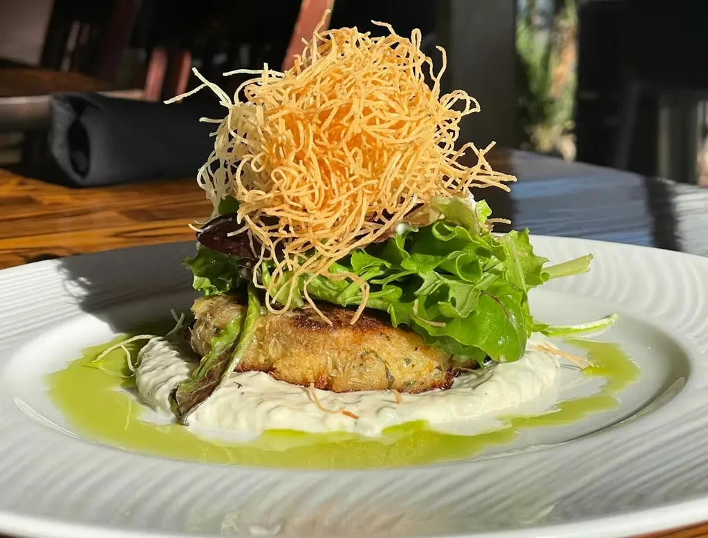 Golden-brown patty on white sauce with green oil, lettuce, fried noodles.