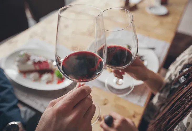 Hands toasting red wine glasses over a berry-topped dessert.
