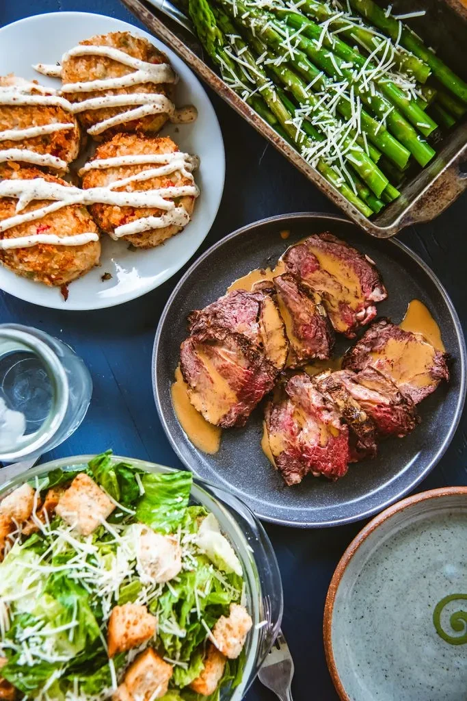 Overhead view of a prepared meal: roast beef, patties, asparagus, salad.