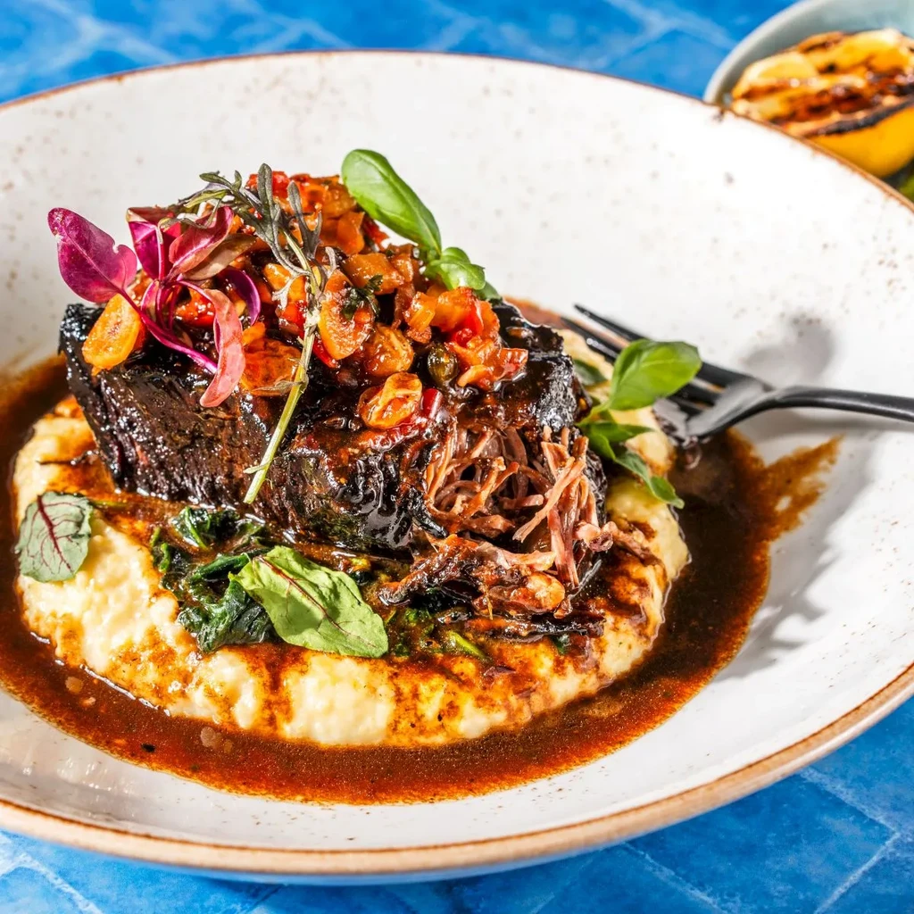 Braised meat, polenta, colorful vegetable relish, and herbs on white plate.