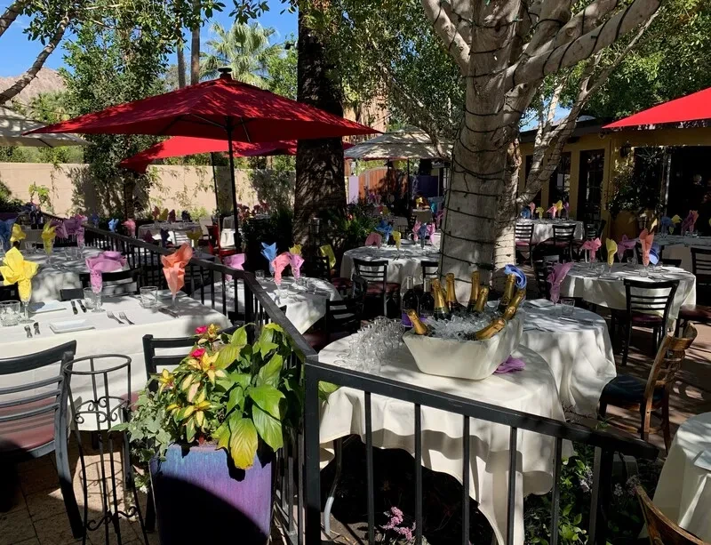Lush outdoor dining patio with white tables, red umbrellas, and champagne.