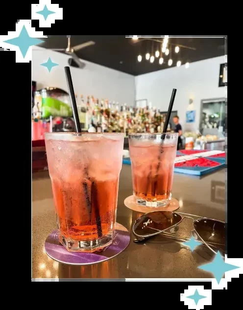Two reddish-pink beverages with ice, straws, and sunglasses on a bar.