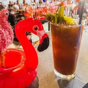 Bloody Mary with garnishes beside a pink flamingo on a bar.